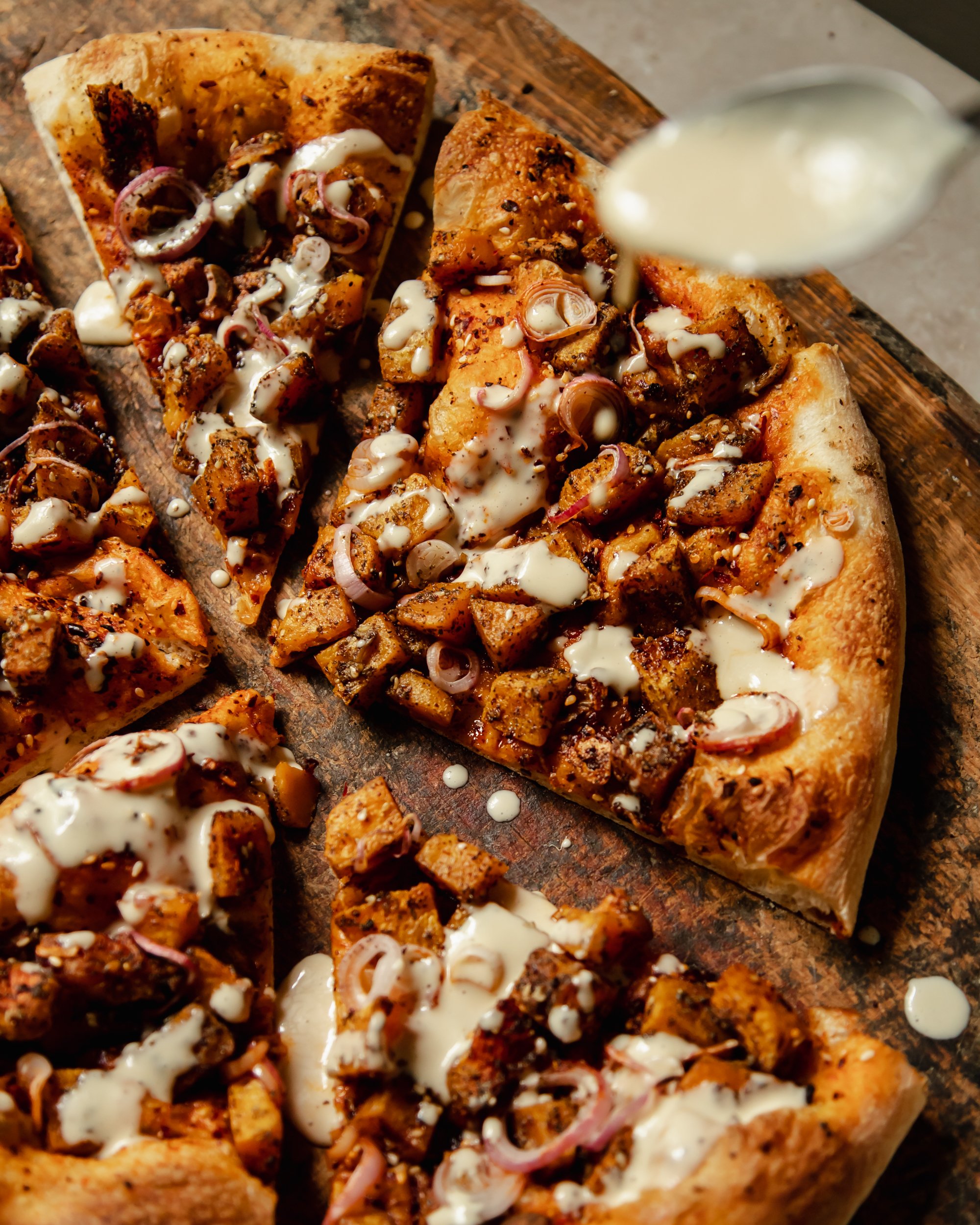 An overhead image shows a butternut squash flatbread being drizzled with a creamy white sauce.