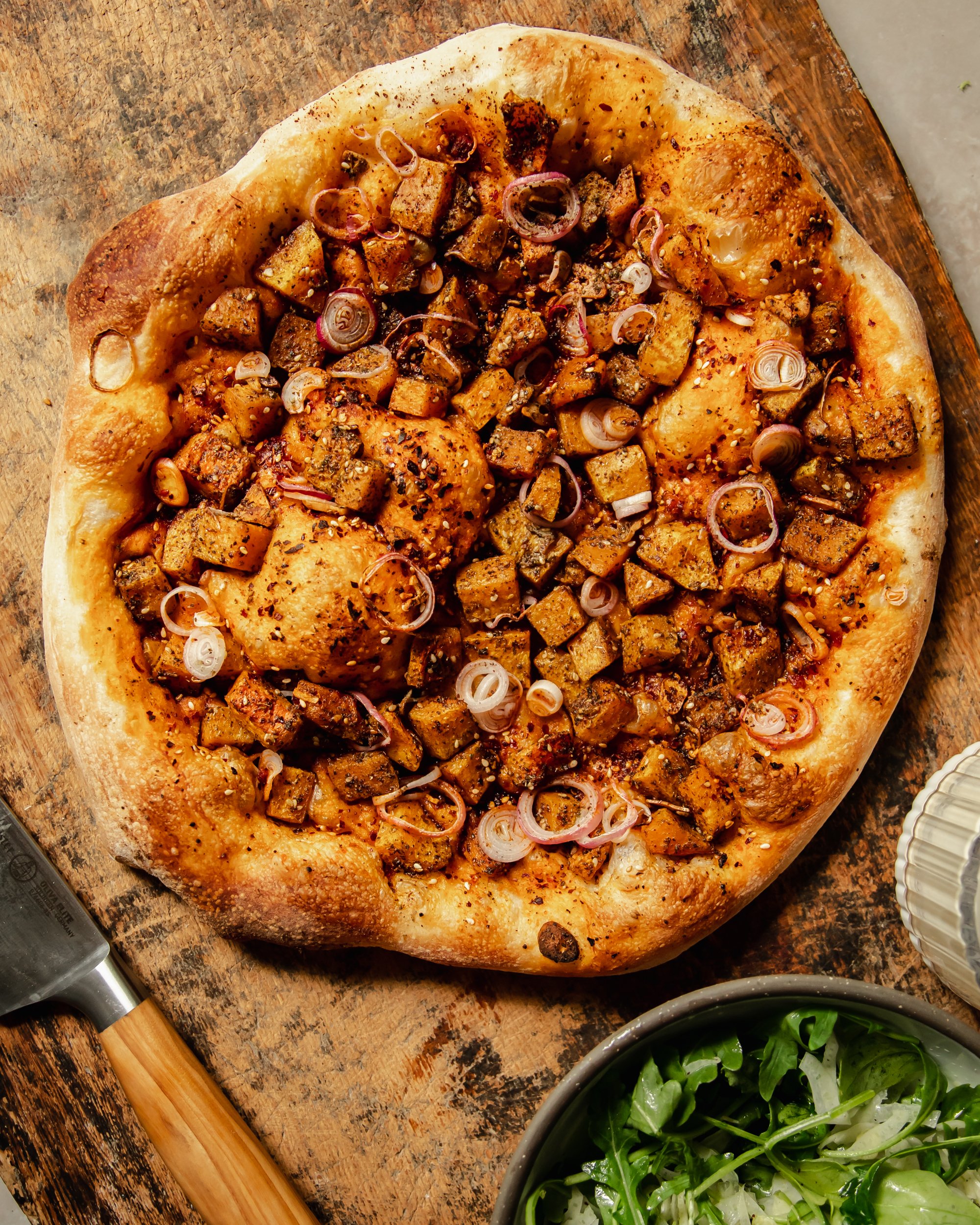 An overhead shot shows a baked butternut squash flatbread before being topped with an arugula fennel salad and a creamy miso garlic sauce.