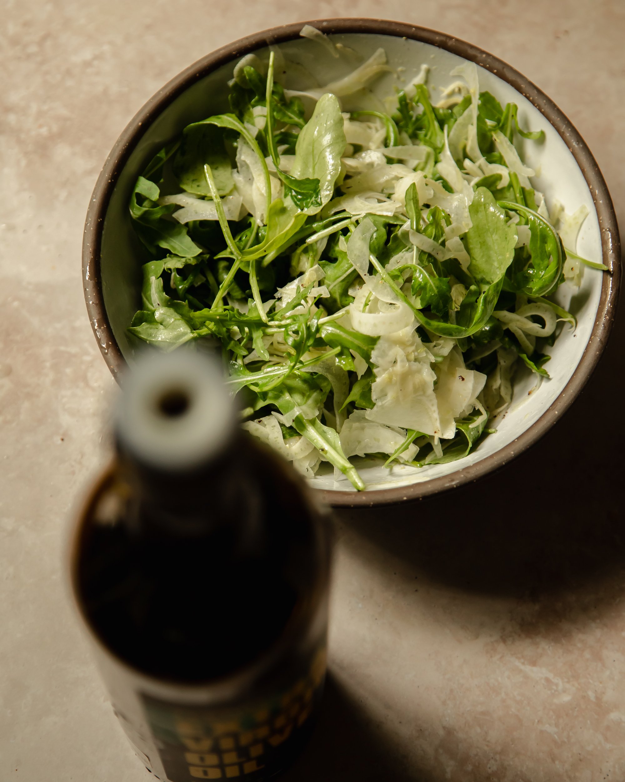 A slight 3/4 angle shot shows a ceramic bowl with a baby arugula and shaved fennel salad. A bottle of olive oil is blurry and in front of the salad bowl.