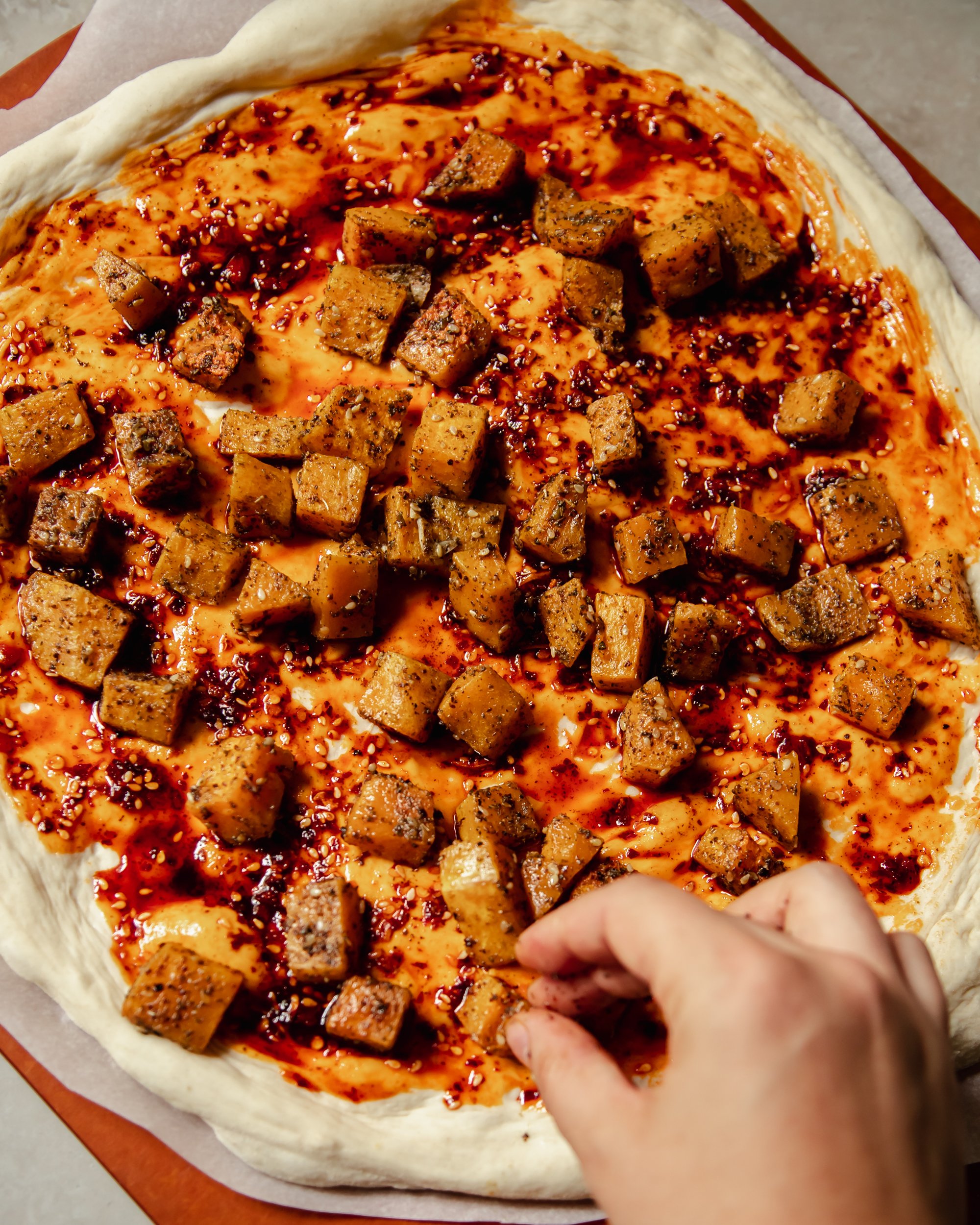 An overhead image shows a flatbread being assembled. Chili oil has been brushed on the surface and roasted cubes of squash are being placed on top by a hand.