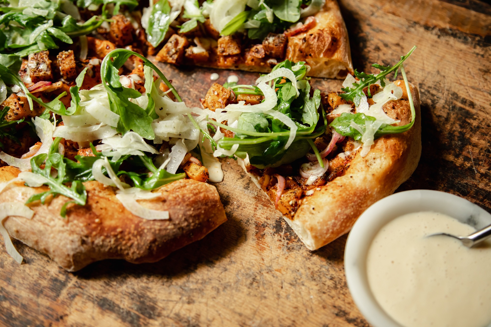 An 3/4 angle shot of a round, vegan butternut squash flatbread topped with an arugula fennel salad and drizzles of a creamy white sauce.