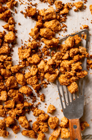 An overhead shot of a baking sheet filled with crispy baked tofu bits. A wooden handled fish spatula is lifting some of the pieces off of the baking sheet.