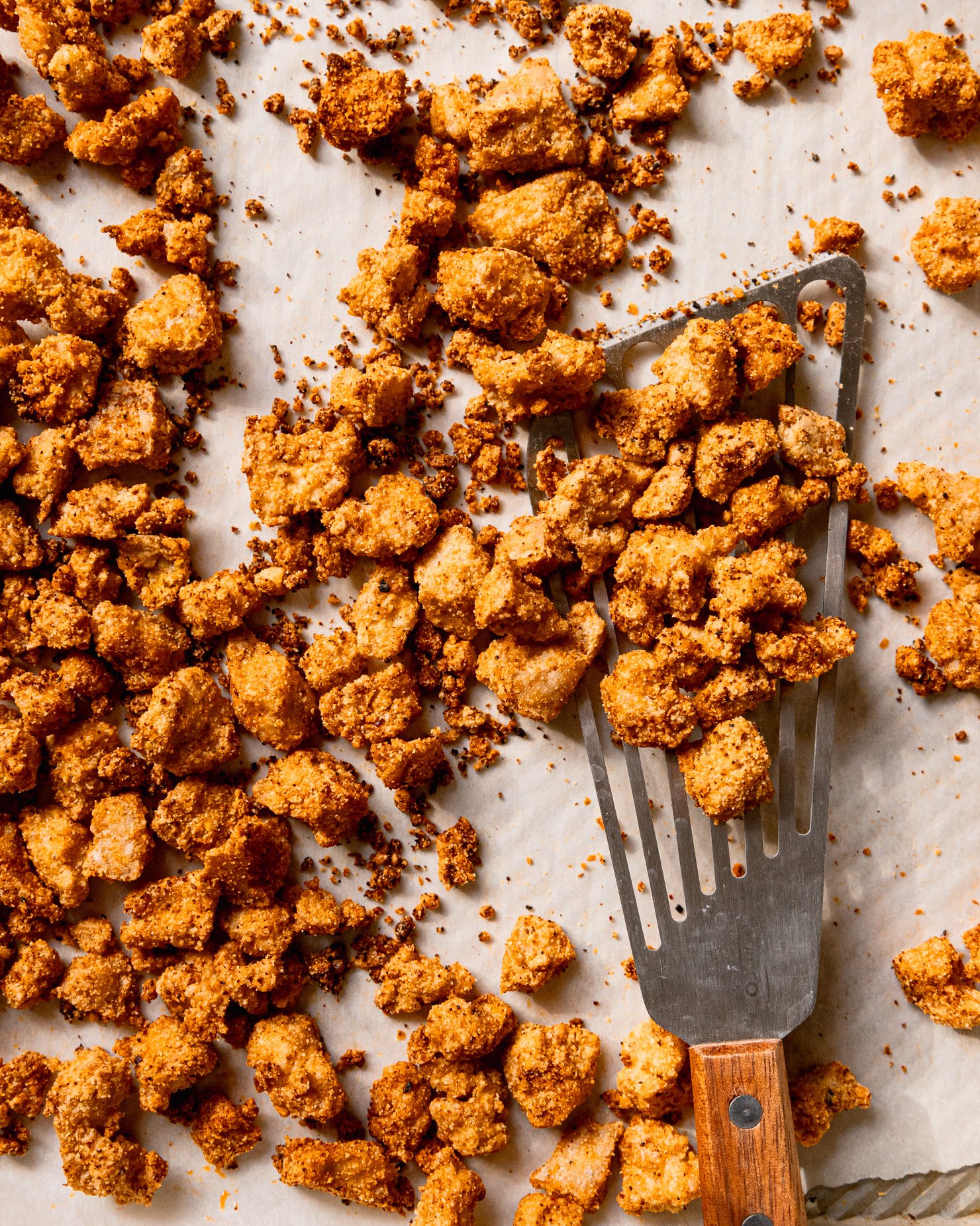 An overhead shot of a baking sheet filled with crispy baked tofu bits. A wooden handled fish spatula is lifting some of the pieces off of the baking sheet.
