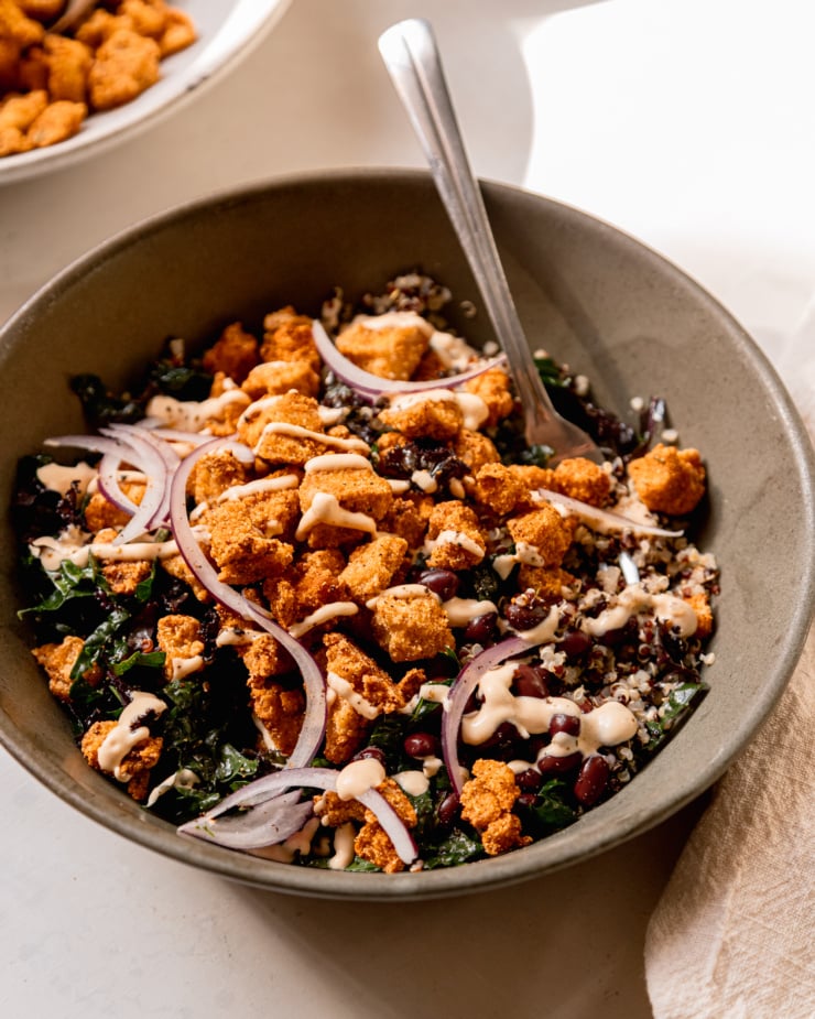 A 3/4 angle shot of a grain bowl with quinoa, kale, black beans, thinly sliced red onion, and crispy baked tofu bits. A creamy sauce is drizzled on top and a fork is sticking out of the bowl.
