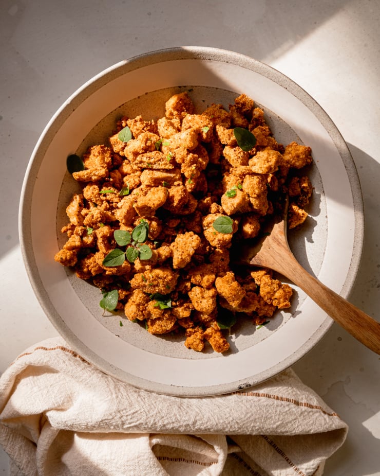 An overhead shot of a light grey bowl filled with crispy nuggets of tofu garnished with fresh herbs. A wooden spoon is sticking out of the serving bowl.