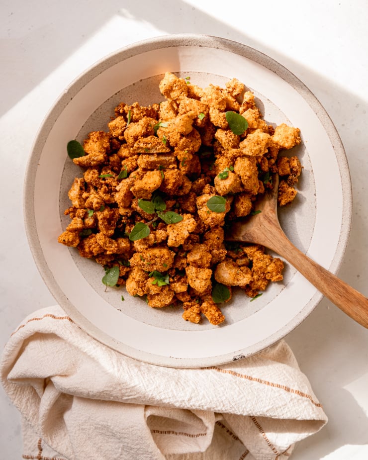 An overhead shot of a light grey bowl filled with crispy nuggets of tofu garnished with fresh herbs. A wooden spoon is sticking out of the serving bowl.