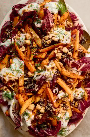 An up close, overhead shot of a harissa carrot and parsnip salad with cooked quinoa, chopped fresh dill, a creamy vegan yogurt sauce dolloped on top, and chopped toasted hazelnuts. All of this is set on a bed of radicchio leaves in a platter