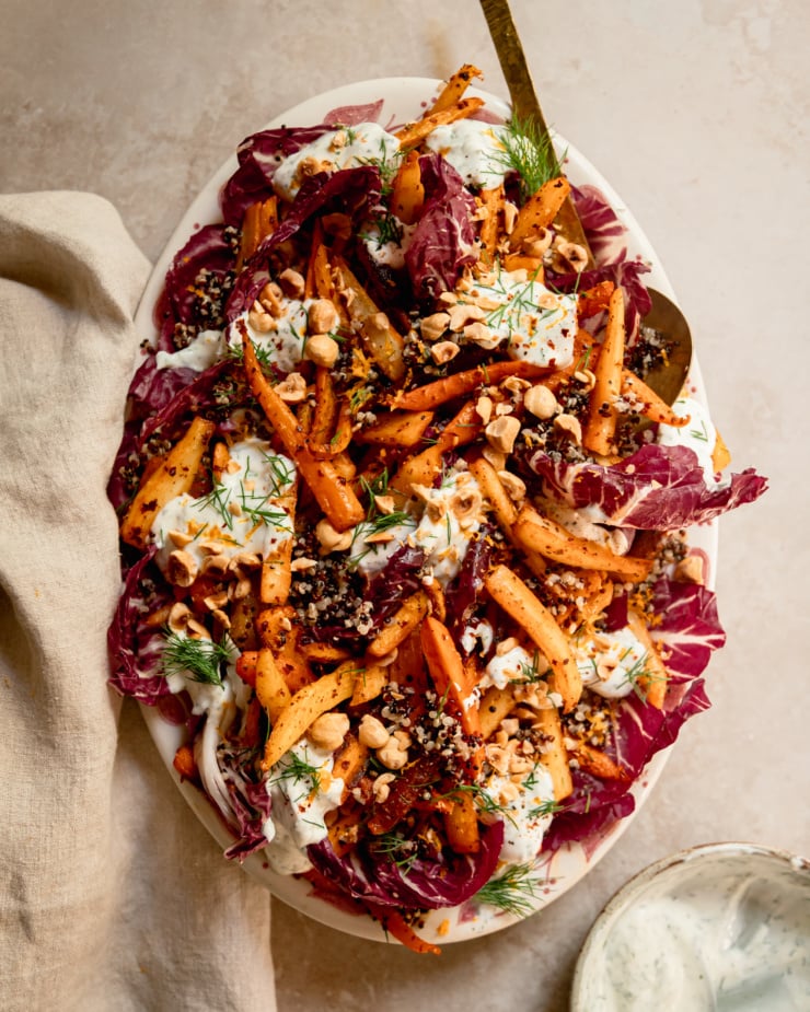 An overhead shot of a harissa carrot and parsnip salad with cooked quinoa, chopped fresh dill, a creamy vegan yogurt sauce dolloped on top, and chopped toasted hazelnuts. All of this is set on a bed of radicchio leaves in a platter