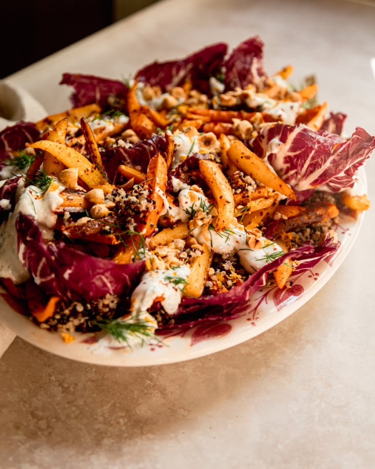 An head on shot of a harissa carrot and parsnip salad with cooked quinoa, chopped fresh dill, a creamy vegan yogurt sauce dolloped on top, and chopped toasted hazelnuts. All of this is set on a bed of radicchio leaves in a platter