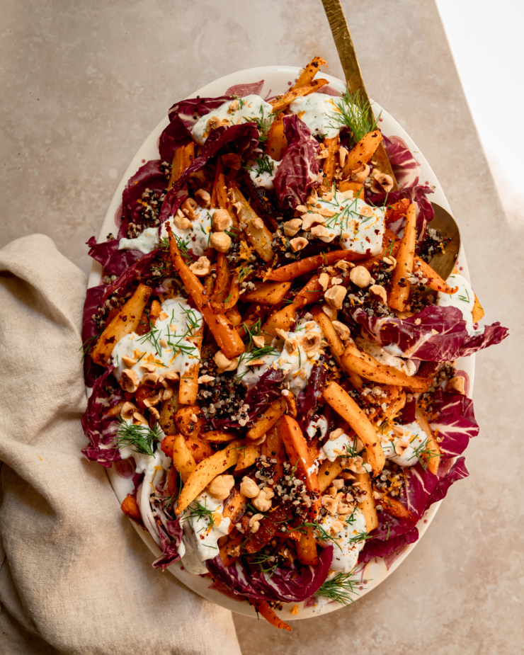 An overhead shot of a harissa carrot and parsnip salad with cooked quinoa, chopped fresh dill, a creamy vegan yogurt sauce dolloped on top, and chopped toasted hazelnuts. All of this is set on a bed of radicchio leaves in a platter