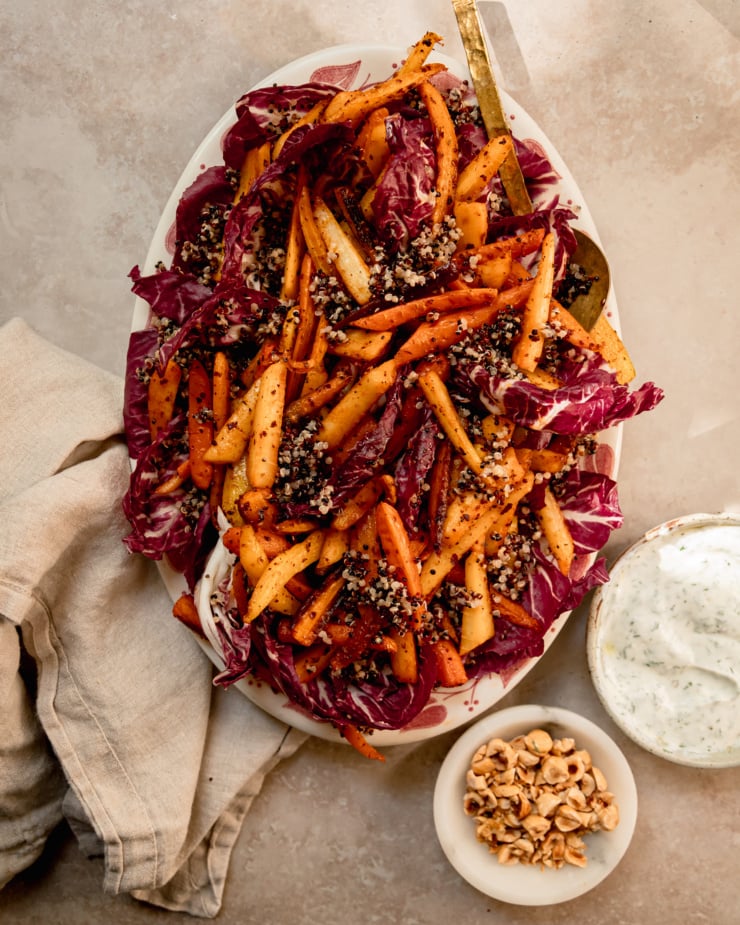 An overhead shot shows a platter with radicchio, cooked quinoa and roasted carrots piled on top. To the side are bowls of vegan yogurt sauce and chopped, toasted hazelnuts.