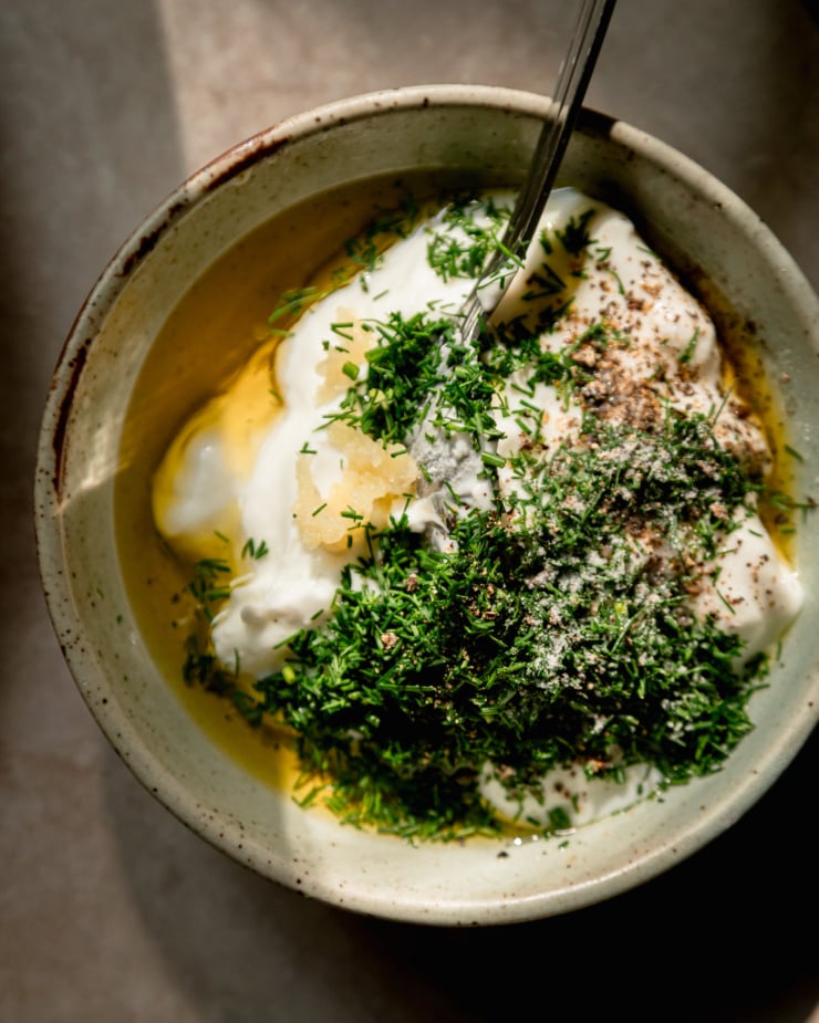 An overhead shot of ingredients for a vegan yogurt sauce in a small grey bowl. Lots of fresh dill is on top and the shot is in direct sunlight.
