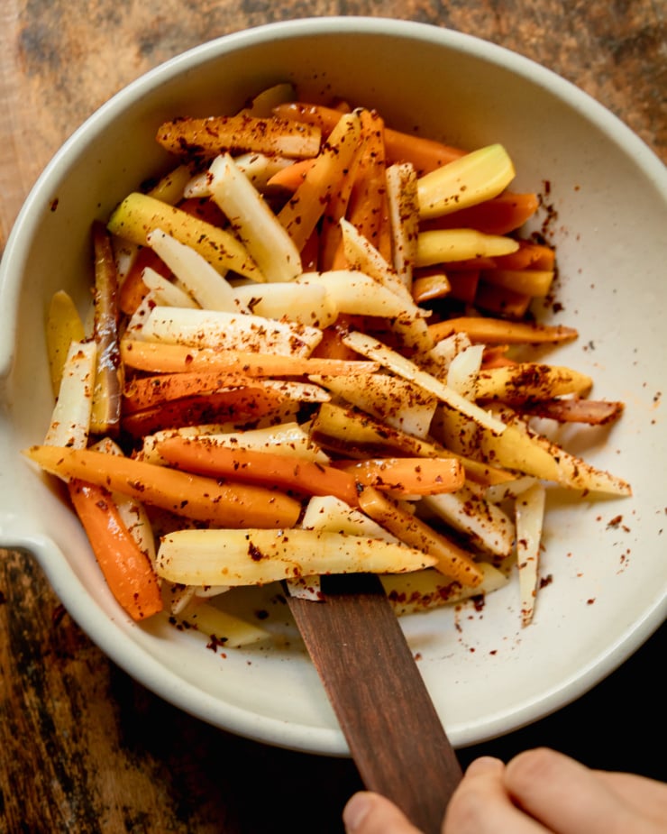 An overhead shot of cut carrots and parsnips being tossed with spices and olive oil in a large bowl.