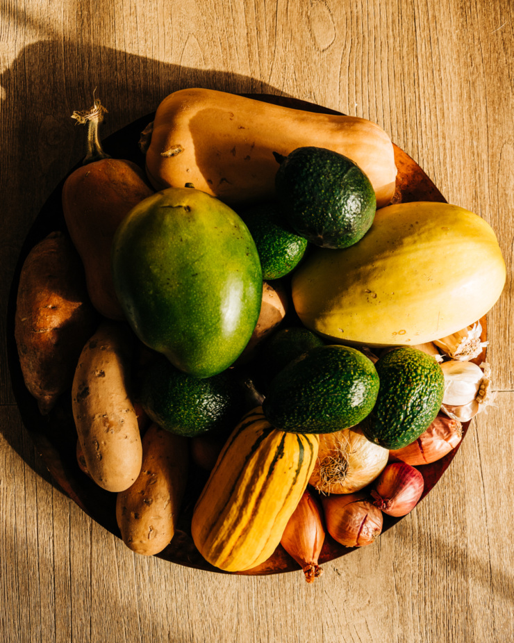 An overhead shot shows a copper tray filled with different types of winter squash, a large mango, avocados, shallots, onions, and garlic.