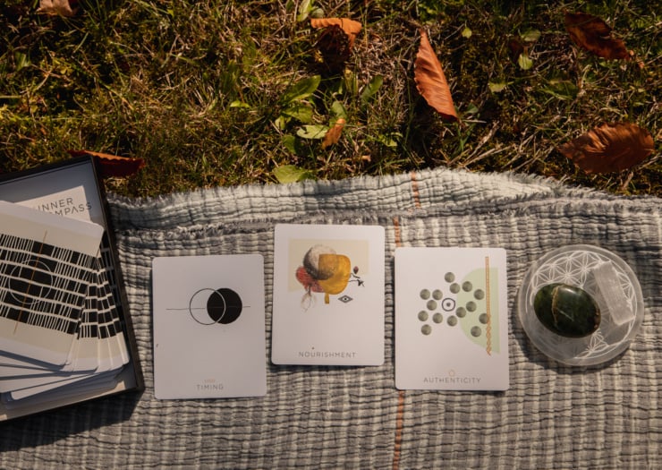 An overhead shot shows 3 Inner Compass oracle cards laid out on a blanket on the grass. There are selenite and jade stones nearby.