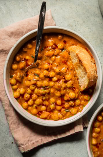 An overhead shot of a bowl of pale orange chickpea soup that is topped with ground pepper, a few rosemary leaves, and drizzles of chili oil. A piece of crusty bread is dunked into the top towards the right of the bowl.