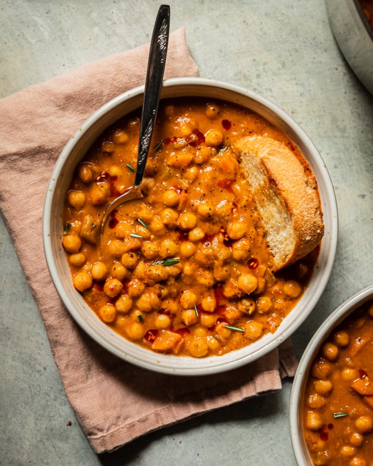 An overhead shot of a bowl of pale orange chickpea soup that is topped with ground pepper, a few rosemary leaves, and drizzles of chili oil. A piece of crusty bread is dunked into the top towards the right of the bowl.