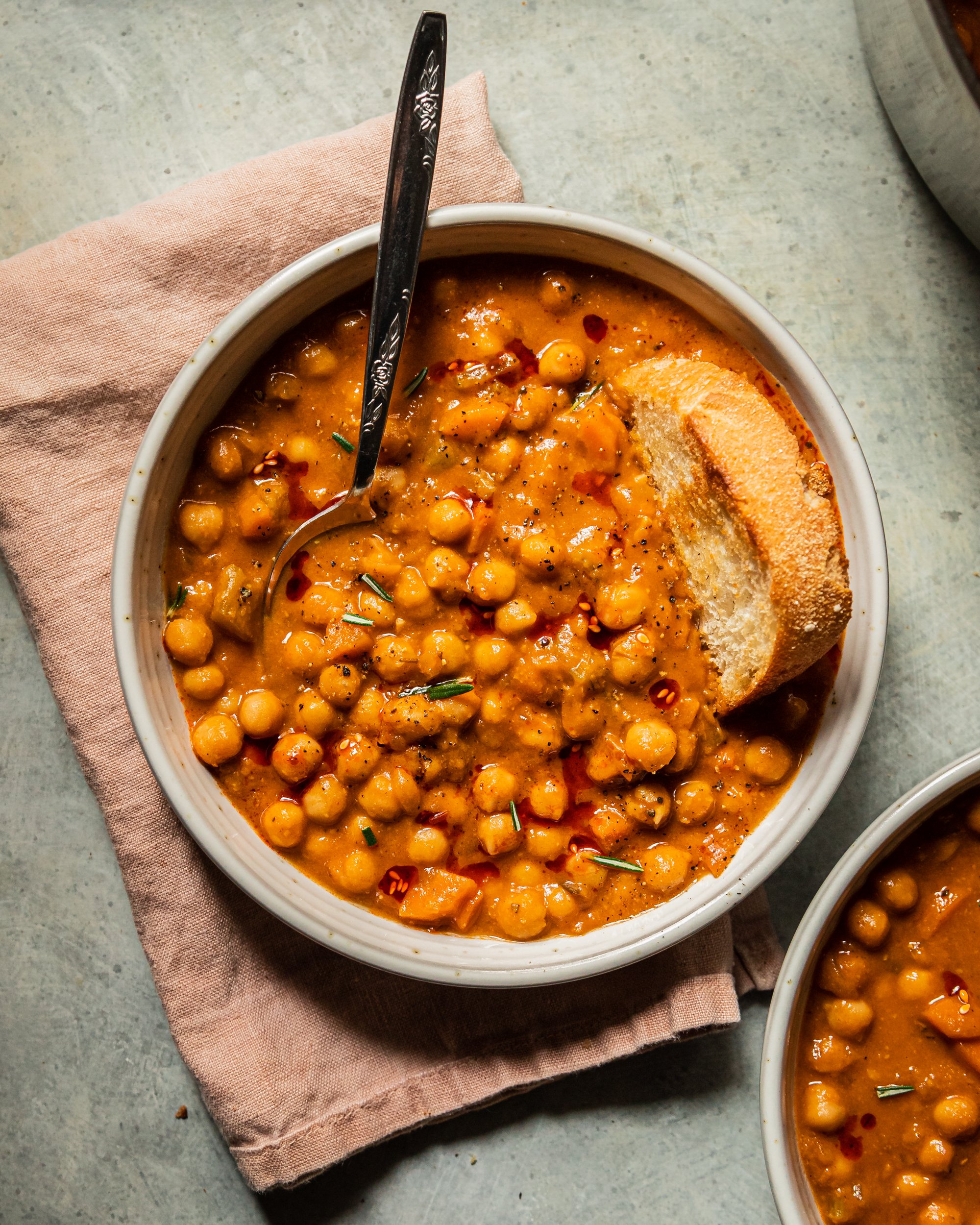 An overhead shot of a bowl of pale orange chickpea soup that is topped with ground pepper, a few rosemary leaves, and drizzles of chili oil. A piece of crusty bread is dunked into the top towards the right of the bowl.