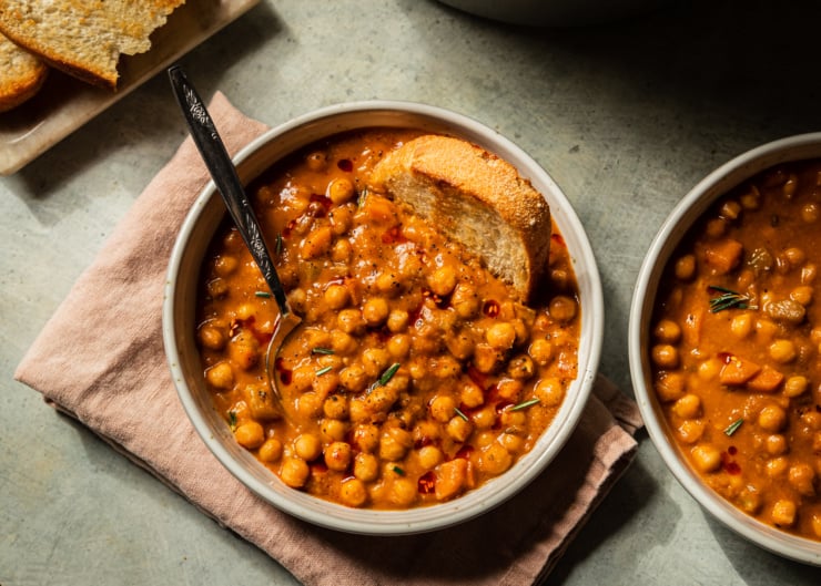 An overhead shot of a bowl of pale orange chickpea soup that is topped with ground pepper, a few rosemary leaves, and drizzles of chili oil. A piece of crusty bread is dunked into the top towards the right of the bowl.