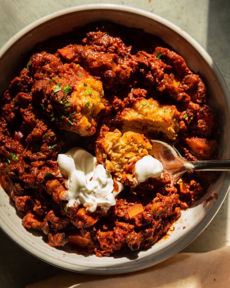 An overhead shot in direct sunlight shows a bowl of vegan chili with cornmeal dumplings and a dollop of vegan sour cream on top. A spoon is sticking out of the bowl.