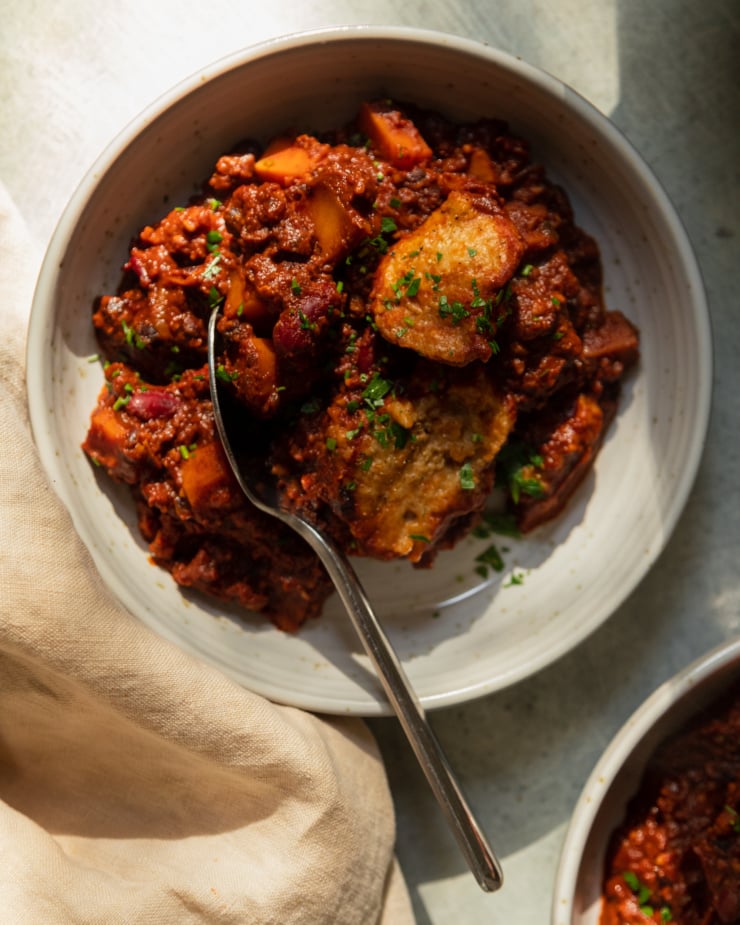 An overhead shot in direct sunlight shows a bowl of vegan chili with a couple of cornmeal dumplings and chopped cilantro on top.