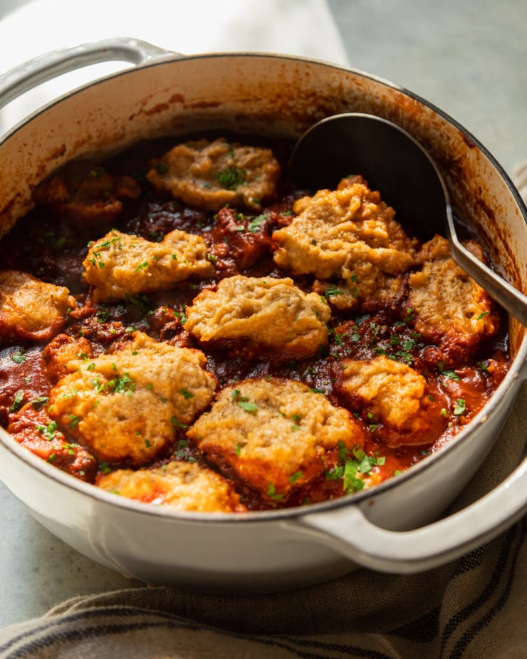 A 3/4 angle shot shows a pot of vegan sweet potato walnut chili in a Dutch oven with puffed up cornmeal dumplings on top. The dish is garnished with chopped parsley and cilantro.