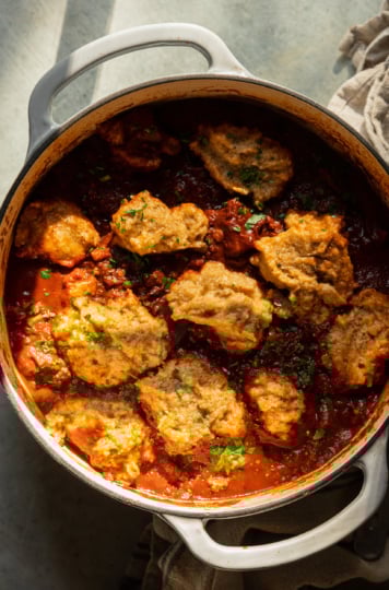 An overhead shot shows a pot of vegan sweet potato walnut chili in a Dutch oven with puffed up cornmeal dumplings on top. The dish is garnished with chopped parsley and cilantro.