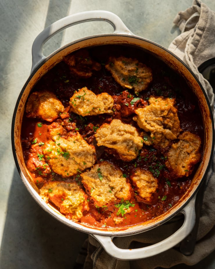 An overhead shot shows a pot of vegan sweet potato walnut chili in a Dutch oven with puffed up cornmeal dumplings on top. The dish is garnished with chopped parsley and cilantro.