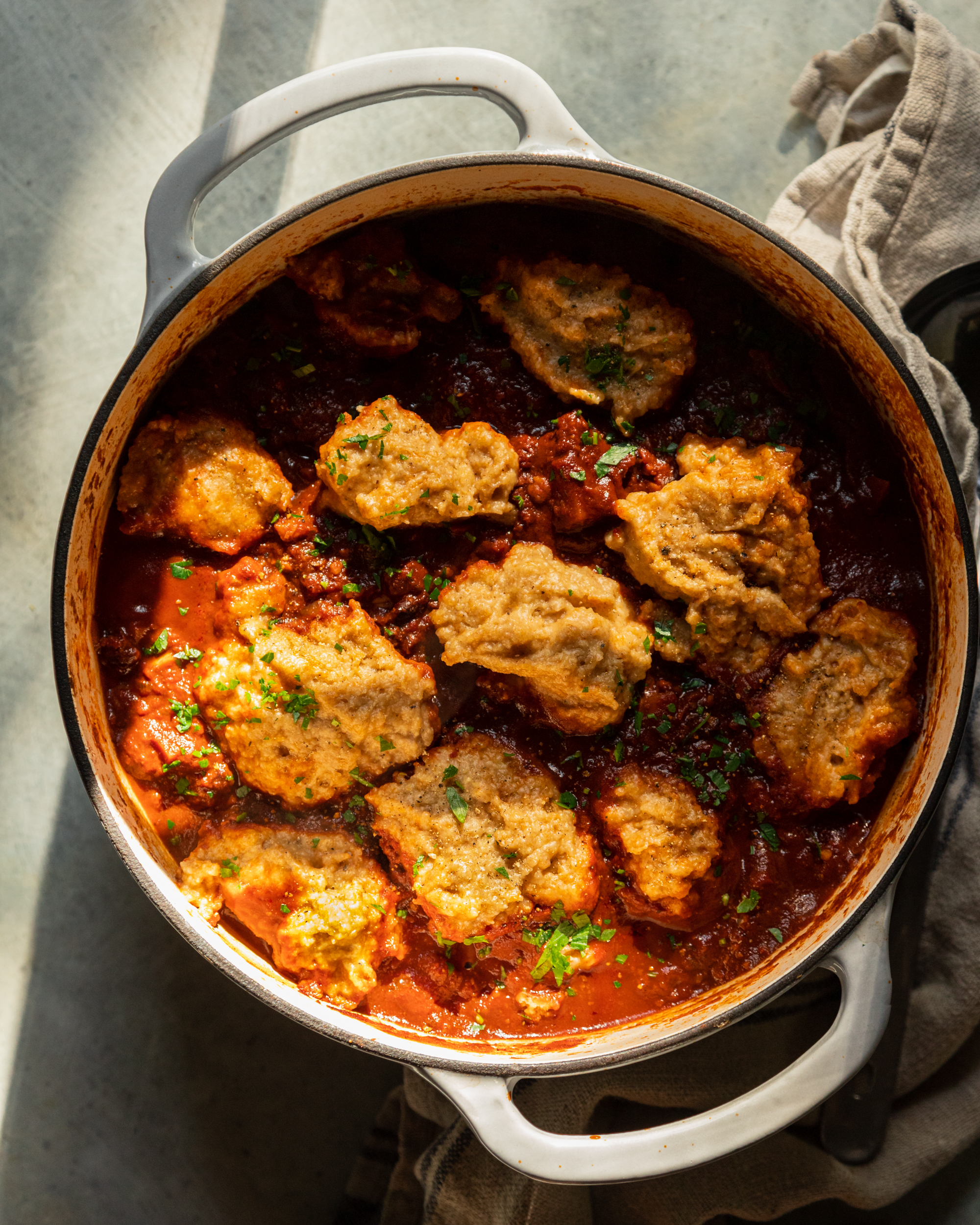 An overhead shot shows a pot of vegan sweet potato walnut chili in a Dutch oven with puffed up cornmeal dumplings on top. The dish is garnished with chopped parsley and cilantro.