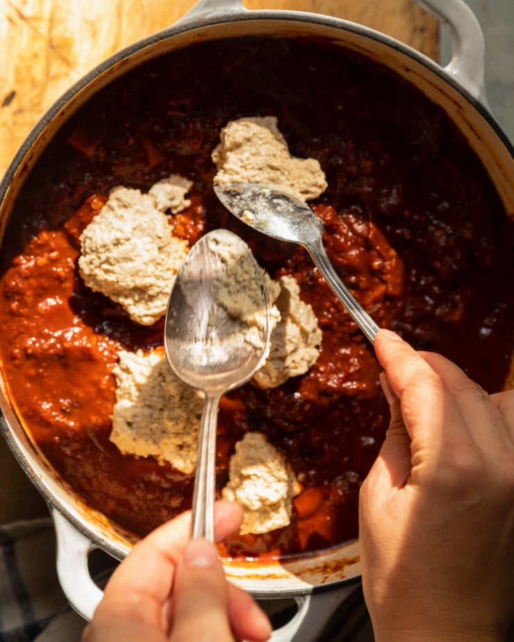 An overhead shot in direct sunlight shows a pair of hands using spoons to dollop cornmeal dumpling dough out on top of a pot of chili.