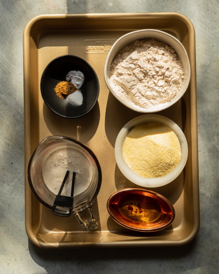 An overhead shot shows a quarter sheetpan with prep for cornmeal dumplings in little bowls and measuring cups.
