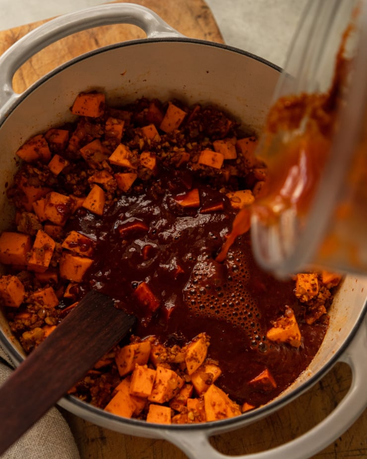 An overhead shot shows a blender pouring in a deep red blended chile mixture into a pot of sweet potatoes and chili base.