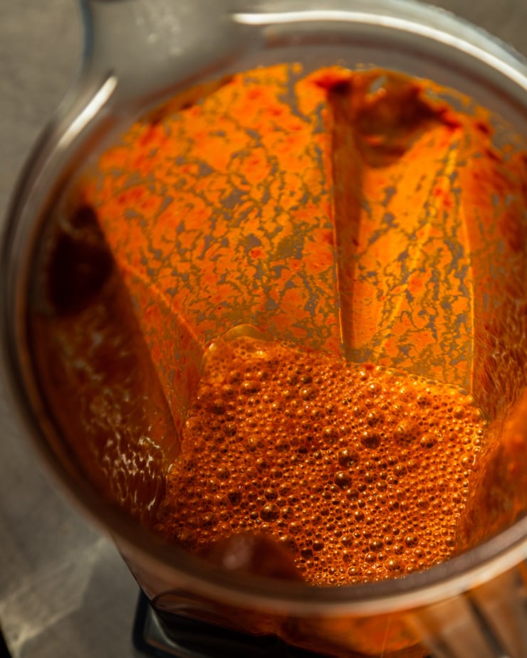 A slight 3/4 angle shot shows a blended chile mixture in a blender pitcher. There are splashes up on the sides of the blender and it appears to be "glowing" in direct sunlight.