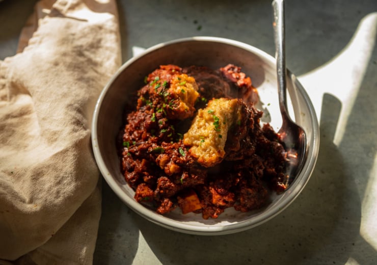 A 3/4 angle shot in harsh light/shadow shows a bowl of vegan chili with a couple of cornmeal dumplings and chopped cilantro on top. There is steam coming off of the bowl.