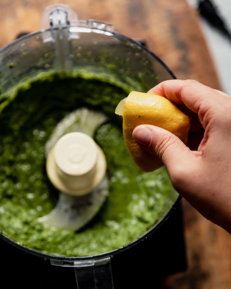 An overhead shot shows a hand squeezing a lemon half into a food processor bowl filled with vegan pesto.