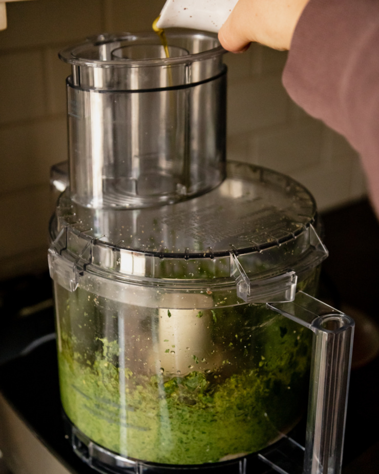 A head-on shot shows a hand pouring a cruet of olive oil into the feed tube of a food processor.