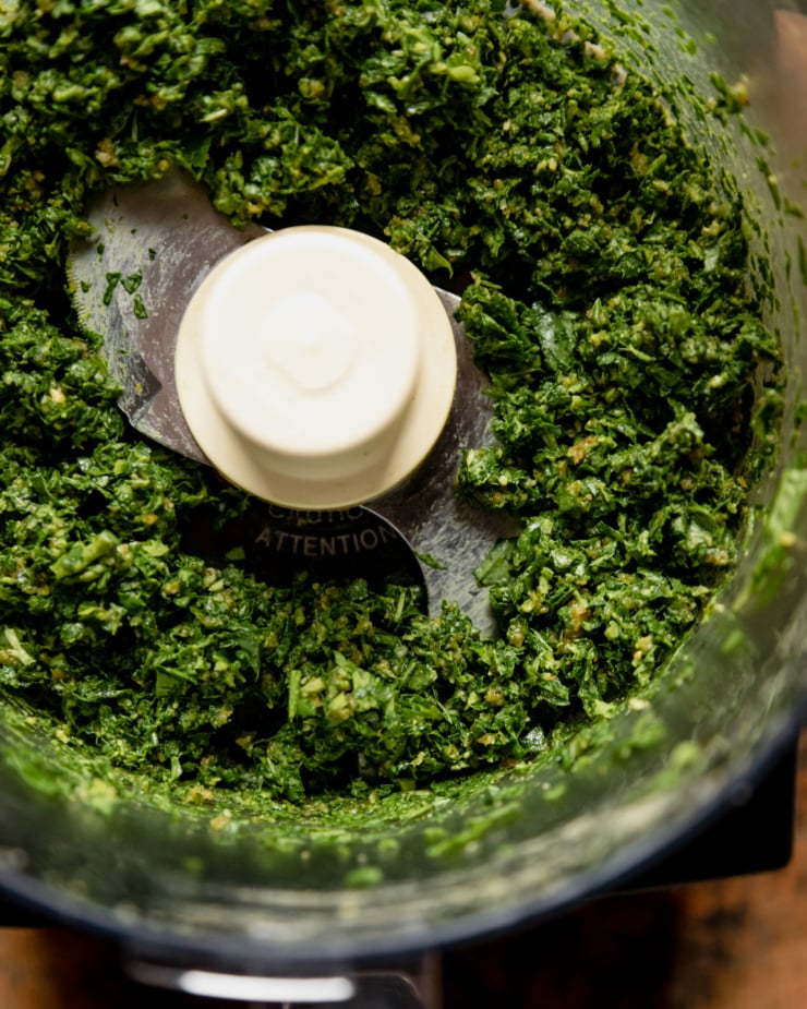 An overhead shot shows a pulsed up mixture of nuts, basil, garlic, and other seasonings.