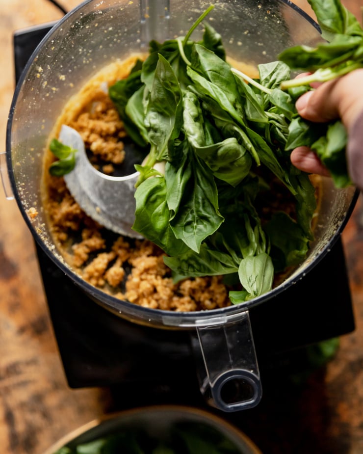 An overhead shot shows a hand tossing fresh basil leaves into a food processor bowl.