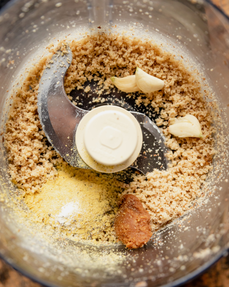 An overhead shot of ground up nuts, garlic cloves, a dab of miso, and nutritional yeast in a food processor.