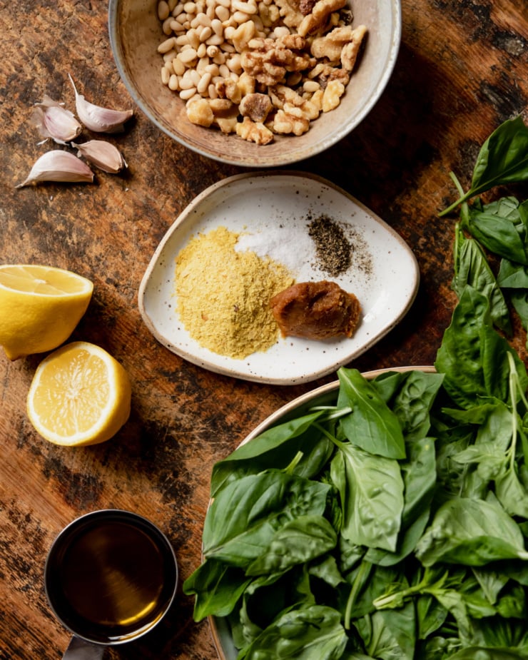 An overhead shot of ingredients for a vegan pesto recipe: walnuts, pine nuts, nutritional yeast, miso, lemon, garlic, olive oil, basil, salt, and pepper.