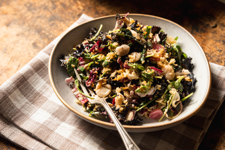 A head on shot of a kale arugula butter bean salad in a wide serving bowl on top of a checkered linen napkin.