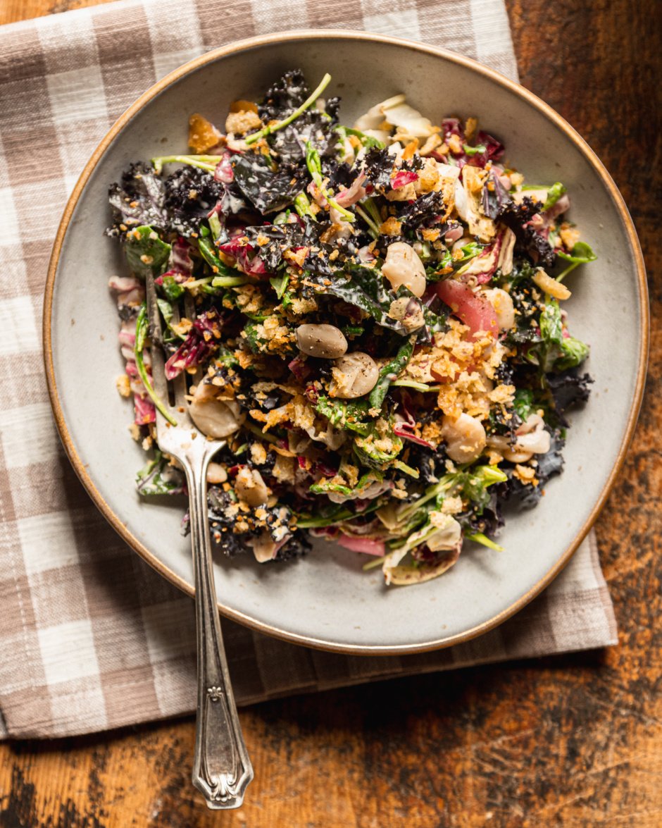 An overhead shot of a kale arugula butter bean salad topped with crispy garlic bread crumbs. The salad is served in a wide serving bowl on top of a linen checkered napkin.