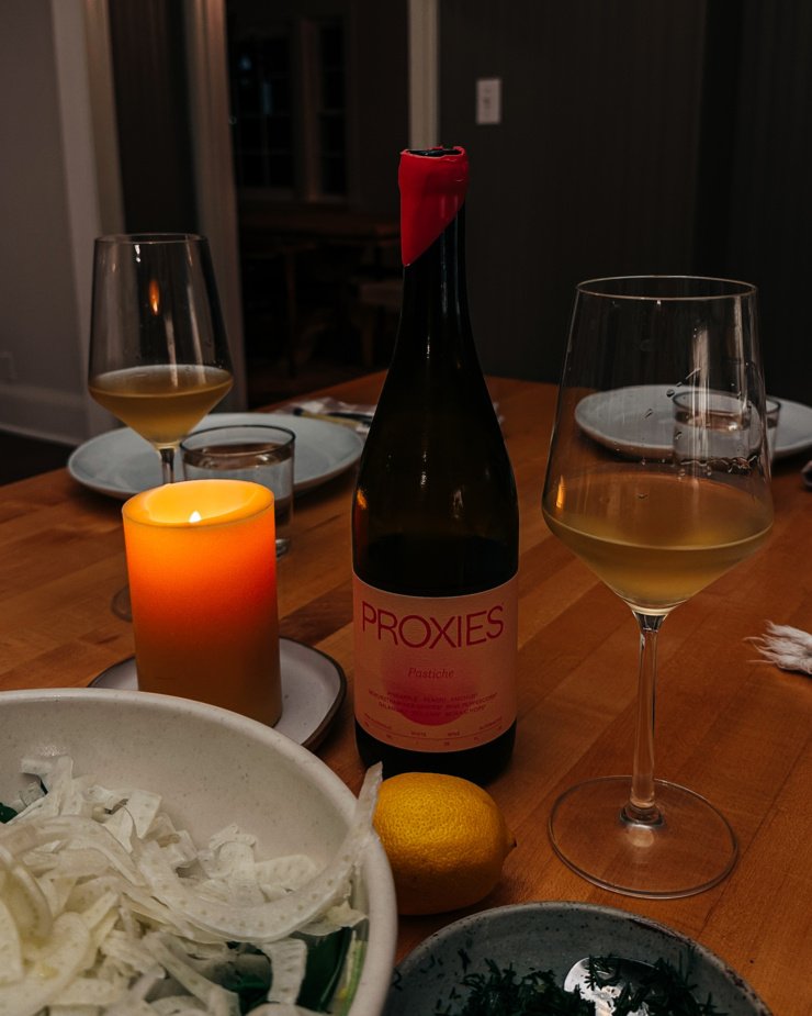 A slight 3/4 angle shot shows a bottle of non-alcoholic wine Proxy with a couple filled wine glasses in dim candlelight.