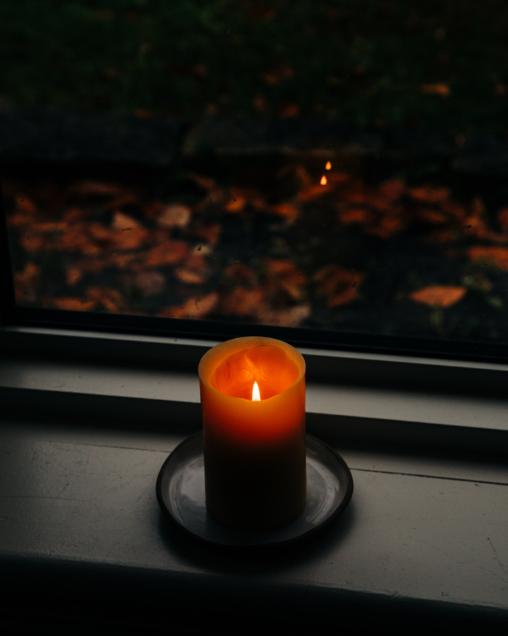 A slight 3/4 angle shot shows a lit candle on a window sill. The lighting is quite dim and autumn leaves can be seen outside the window.