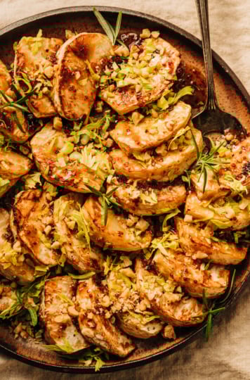 An overhead shot of cider glazed celery root pieces fanned out on a speckled brown plate. The celery root is topped with chopped walnuts, rosemary leaves, and finely chopped celery heart leaves.
