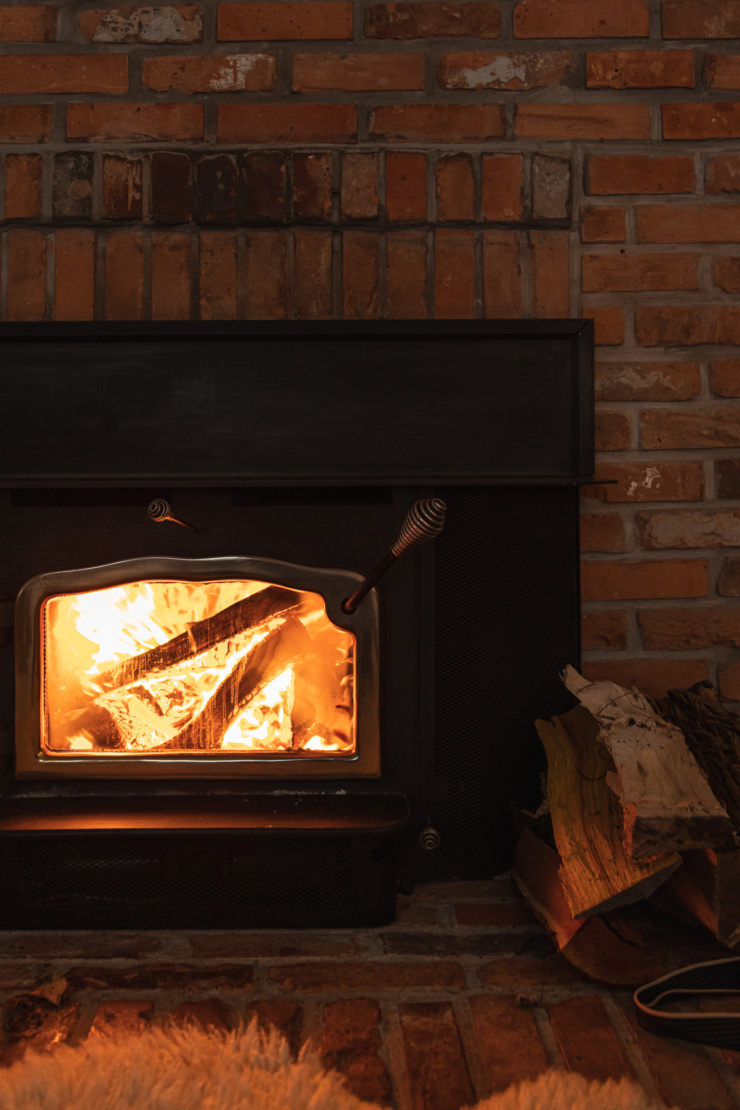 A head on shot of a fire burning in a wood stove set against a pink-ish red brick fireplace. A stack of chopped wood is nearby.