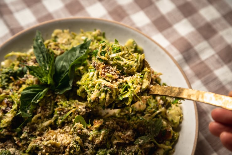 A 3/4 angle, up close shot of sautéed shaved brussels sprouts in a shallow bowl, topped with balsamic glaze, fresh basil and vegan "parmesan." A brass spoon is lifting some of the vegetable side dish out of the bowl.