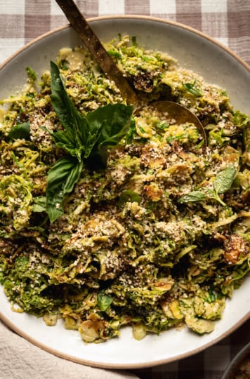 An overhead shot of sautéed shaved brussels sprouts in a shallow bowl, topped with balsamic glaze, fresh basil and vegan "parmesan"