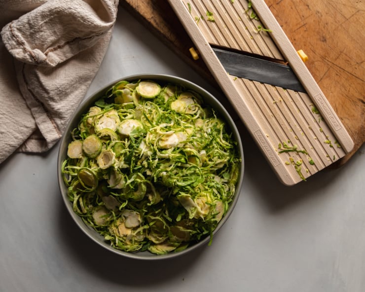 An overhead shot shows a mandoline slicer and some shredded brussels sprouts in a wide bowl.