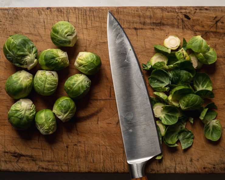 An overhead shot shows a cutting board with trimmed brussels sprouts and a chef's knife. The discarded trimmings are to the side.